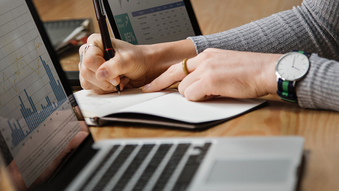 Accountant taking handwritten notes beside a laptop showing financial charts and a spreadsheet during a quality of earnings review